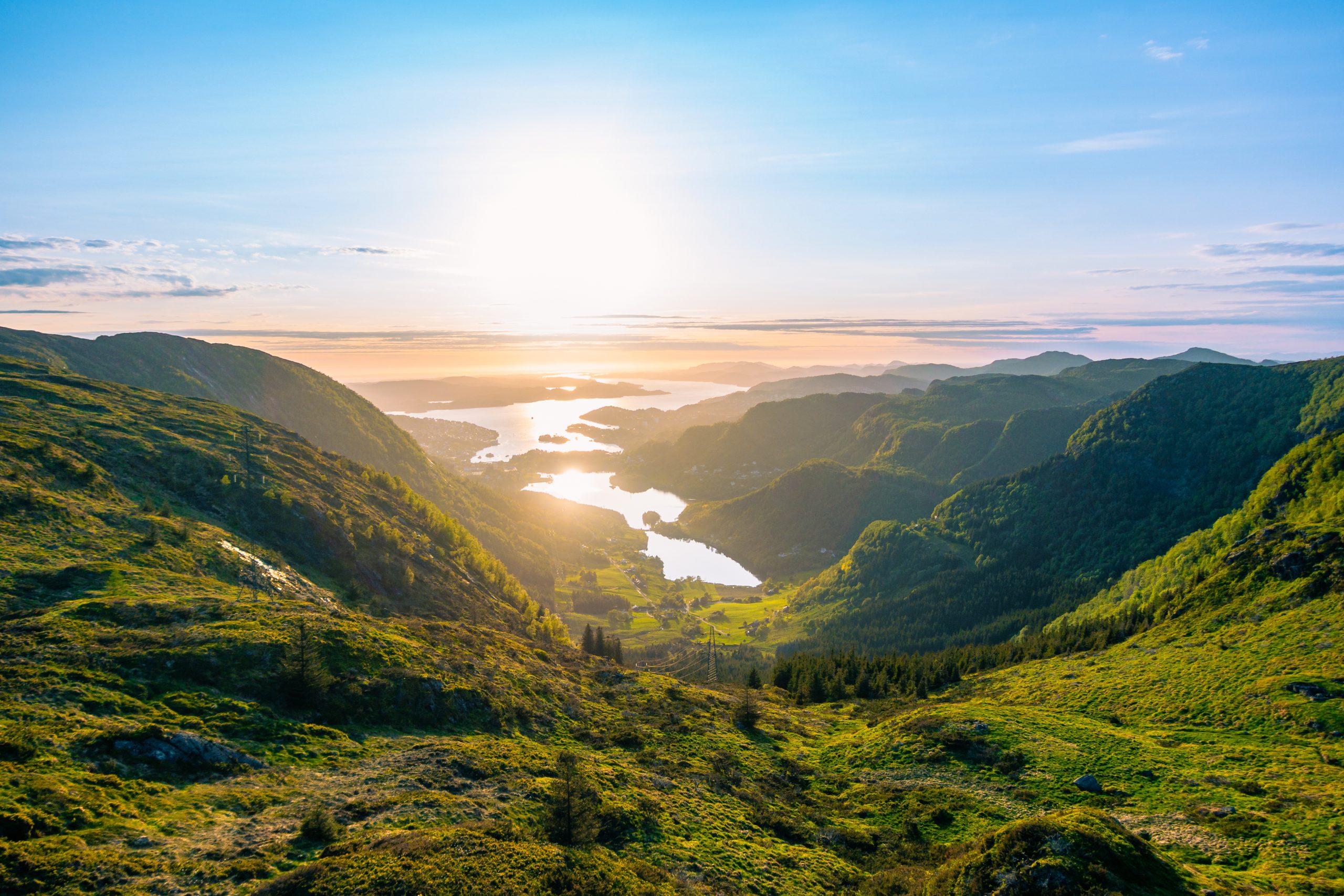 Lush green tree, grass and marsh lined valley leading into the fjord surrounding the nordic city Bergen, Norway, at the start of a midnight sunset with an overpoweringly strong sun
